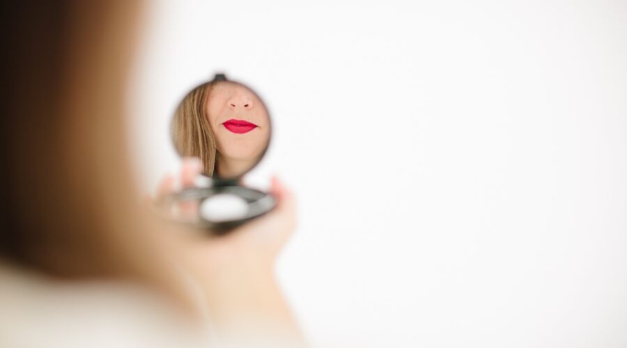 In a diffuse image we can see a round makeup mirror, where the area of ​​the mouth and chin of a light brown woman is reflected.